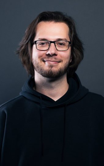 Armend Bajramaj, ein Mann mit schulterlangem braunem Haar und Brille, lächelt leicht. Er trägt einen schwarzen Kapuzenpullover und steht vor einem schlichten dunkelgrauen Hintergrund.