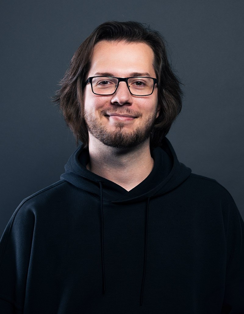 Armend Bajramaj, ein Mann mit schulterlangem braunem Haar und Brille, lächelt leicht. Er trägt einen schwarzen Kapuzenpullover und steht vor einem schlichten dunkelgrauen Hintergrund.