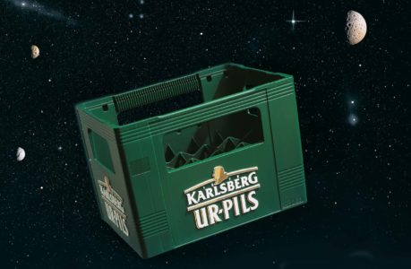 Eine grüne Karlsberg UrPils-Bierkiste schwebt im Weltall, umgeben von Sternen und fernen Planeten, und zelebriert die Abenteuerlust von Bierliebhabern überall.