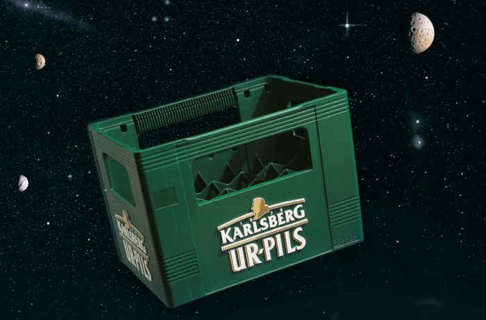 Eine grüne Karlsberg UrPils-Bierkiste schwebt im Weltall, umgeben von Sternen und fernen Planeten, und zelebriert die Abenteuerlust von Bierliebhabern überall.
