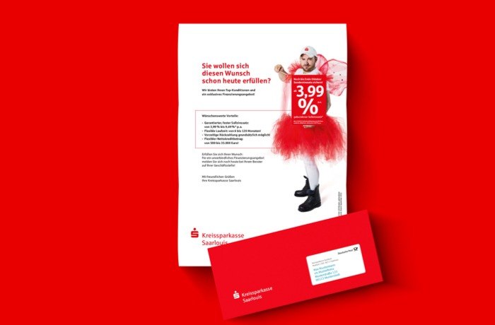 Eine Broschüre und ein roter Umschlag der KSK Saarlouis auf rotem Hintergrund. Die Broschüre zeigt ein Kind in einem Feenkostüm und wirbt für einen Privatkredit mit einem Zinssatz von 3,99 %. Text und Logos sind in deutscher Sprache.
