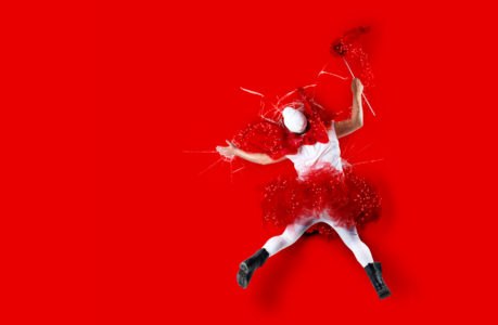 Eine Person in rotem Tutu, weißem Hemd, weißer Strumpfhose, schwarzen Stiefeln und weißer Maske liegt auf einem leuchtend roten Hintergrund und hält einen rot-weißen Zauberstab mit rotem Pulver in der Hand, als würde sie aus den Träumen der KSK Saarlouis Privatkredit zaubern.