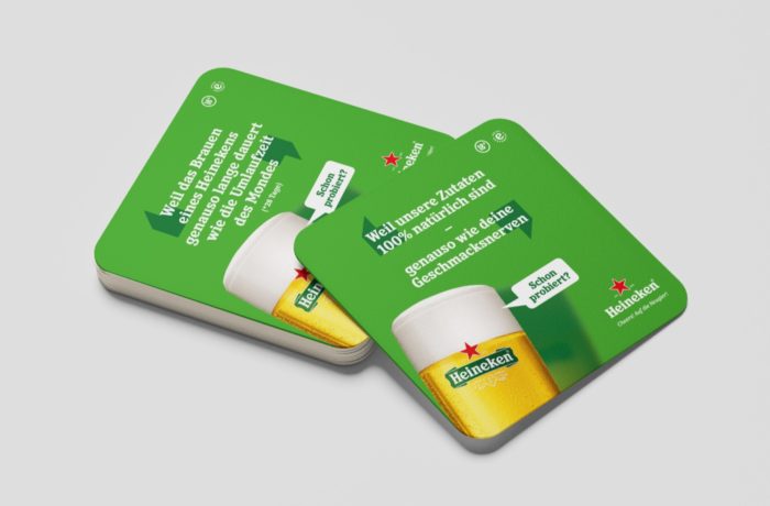 Grüne Heineken-Bieruntersetzer mit einem Glas Bier und einem deutschen Text über 100% natürliche Zutaten. Die Untersetzer zeigen das Heineken-Logo und einen weißen Stern auf der Oberseite.