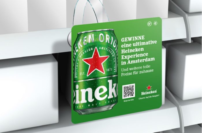 Eine grüne Heineken-Dose ist auf einer hängenden Display-Karte in einem Ladengang abgebildet. Die Karte ist auf Deutsch und wirbt für ein Gewinnspiel, bei dem man ein Heineken-Erlebnis in Amsterdam gewinnen kann.