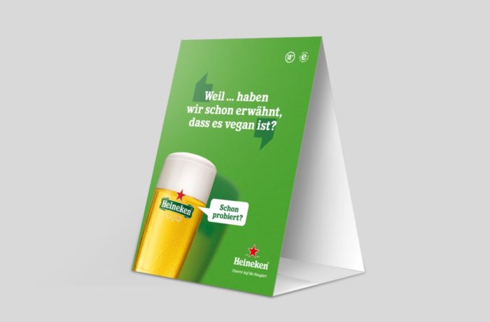 Eine grüne Heineken-Tischkarte mit einem Glas Bier, dem Heineken-Logo und einem deutschen Text, der darauf hinweist, dass Heineken vegan ist. Der Hintergrund ist einfarbig grau.
