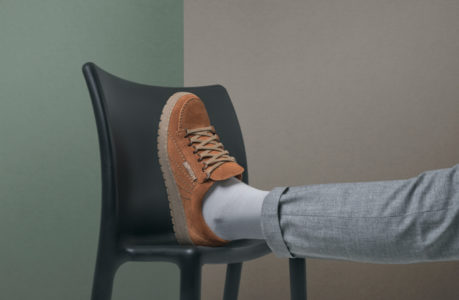 Eine Person, die eine hellgraue Hose und weiße Socken trägt, ruht mit ihrem Fuß in einem braunen Wildleder-Sneaker auf der Sitzfläche eines schwarzen Stuhls vor einem zweifarbigen Hintergrund in Grün und Beige.