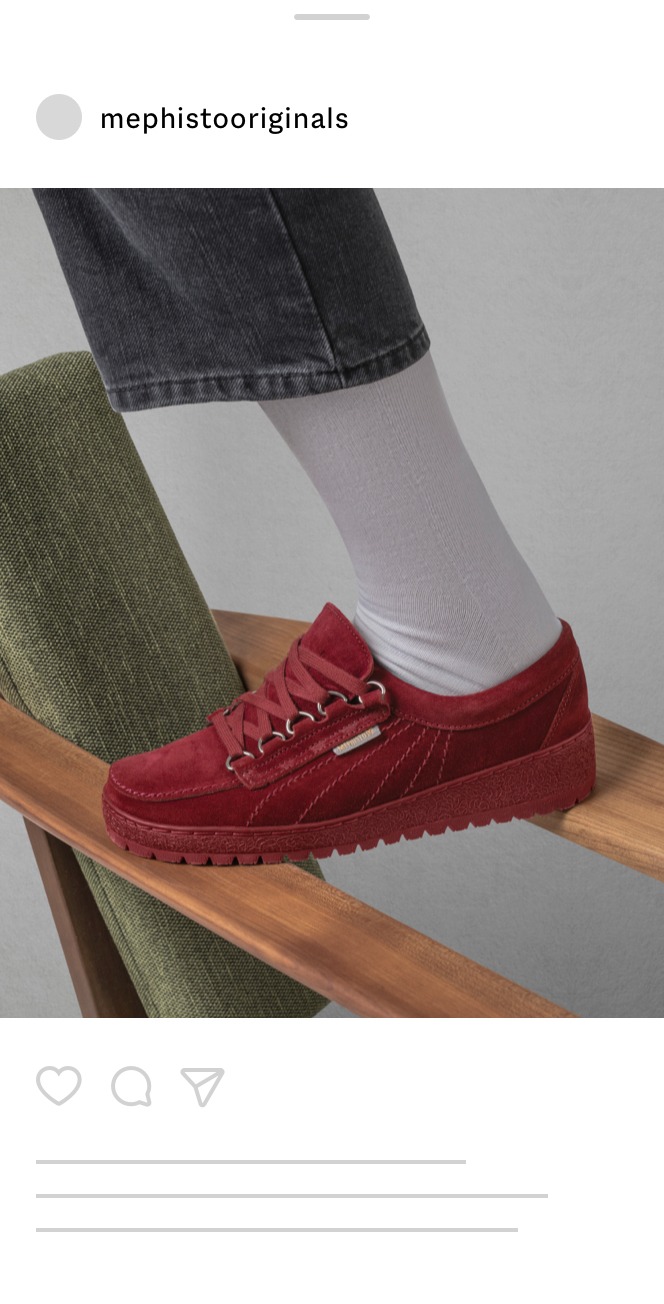 Eine Person, die weiße Socken und rote Mephisto Originals-Wildlederschuhe trägt, steht mit einem Fuß auf einem Holzstuhl und mit dem anderen auf einem grün gepolsterten Stuhl. Das Bild ist wie ein Social-Media-Beitrag gestylt.