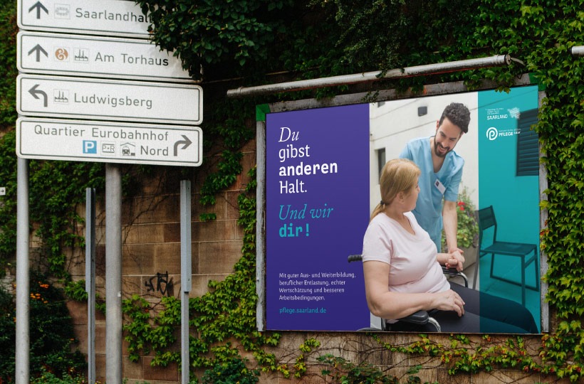 Eine Plakatwand zeigt einen Krankenpfleger, der lächelnd mit einer Frau im Rollstuhl spricht. Der deutsche Text, "Du gibst anderen Halt. Und wir dir!" wirbt für Gesundheitskarrieren des Ministeriums für Gesundheit. In der Nähe sind Straßenschilder zu sehen.