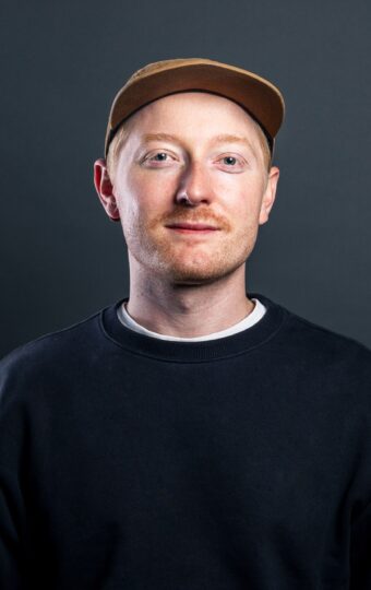 Benjamin Mordiconi, der eine hellbraune Mütze, ein schwarzes Sweatshirt und ein weißes Hemd trägt, steht vor einem dunkelgrauen Hintergrund und schaut mit neutralem Blick in die Kamera.