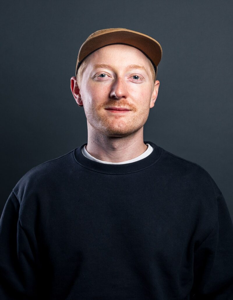 Benjamin Mordiconi, der eine hellbraune Mütze, ein schwarzes Sweatshirt und ein weißes Hemd trägt, steht vor einem dunkelgrauen Hintergrund und schaut mit neutralem Blick in die Kamera.