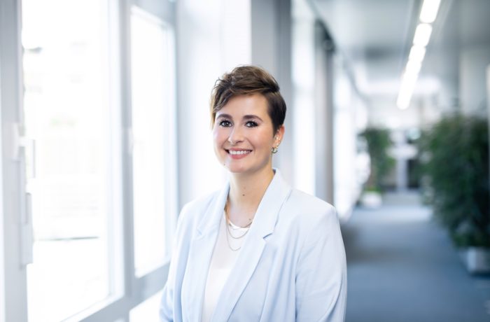 Eine Person mit kurzen braunen Haaren, die einen hellblauen Blazer und ein weißes Oberteil trägt, lächelt, während sie in einem hellen Flur mit Fenstern und Pflanzen im Hintergrund steht. Eine Person mit kurzen braunen Haaren, die einen hellblauen Blazer und ein weißes Oberteil trägt, lächelt, während sie in einem hellen Flur mit Fenstern und Pflanzen im Hintergrund steht.