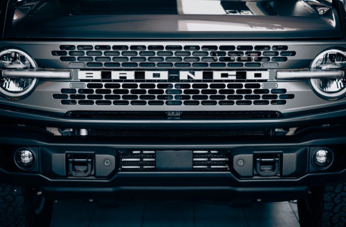 Nahaufnahme des Kühlergrills eines dunkel gefärbten Ford Bronco mit dem markanten BRONCO-Schriftzug, den runden Scheinwerfern und den robusten Stoßfängerdetails. Nahaufnahme des Kühlergrills eines dunkel gefärbten Ford Bronco mit dem markanten BRONCO-Schriftzug, den runden Scheinwerfern und den robusten Stoßfängerdetails.
