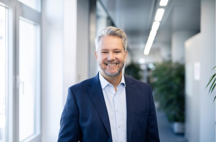 Ein lächelnder Mann mit grauem Haar und Bart, der einen blauen Anzug und ein hellblaues Hemd trägt, steht in einem modernen Büroflur mit großen Fenstern und Grünflächen im Hintergrund. Ein lächelnder Mann mit grauem Haar und Bart, der einen blauen Anzug und ein hellblaues Hemd trägt, steht in einem modernen Büroflur mit großen Fenstern und Grünflächen im Hintergrund.