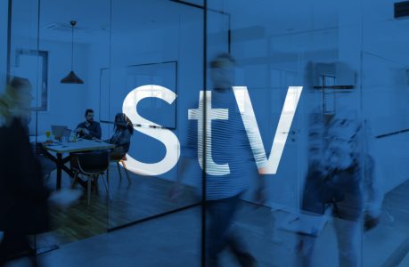 Büroszene mit verschwommenen Personen, die an Glaswänden mit großen, weißen Buchstaben StV vorbeigehen. Im Hintergrund sitzt eine Gruppe um einen Tisch in einem Besprechungsraum. Das Gesamtbild ist blau eingefärbt.
