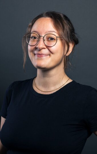 Malika Hagemann, eine Frau mit Brille und zurückgebundenen braunen Haaren, steht lächelnd vor einem dunklen Hintergrund. Sie trägt ein schwarzes T-Shirt und hat eine Hand in die Hüfte gestemmt.