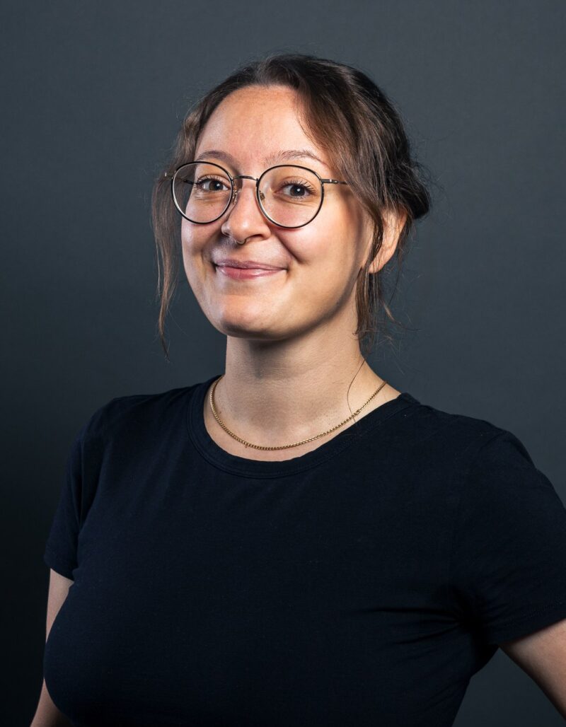 Malika Hagemann, eine Frau mit Brille und zurückgebundenen braunen Haaren, steht lächelnd vor einem dunklen Hintergrund. Sie trägt ein schwarzes T-Shirt und hat eine Hand in die Hüfte gestemmt.