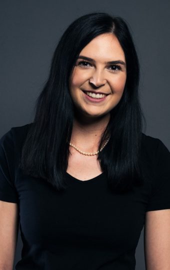 Mia Roepnack, eine Frau mit langen dunklen Haaren, trägt ein schwarzes kurzärmeliges Oberteil und eine beigefarbene Hose, während sie lächelnd vor einem dunkelgrauen Hintergrund steht.