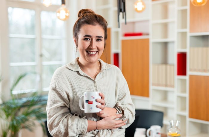 Eine Frau mit braunem Haar in einem Dutt und einem hellen Oberteil lächelt, während sie eine weiße Tasse in einem hellen, modernen Büro mit Regalen, Büchern und warmer Beleuchtung im Hintergrund hält. Eine Frau mit braunem Haar in einem Dutt und einem hellen Oberteil lächelt, während sie eine weiße Tasse in einem hellen, modernen Büro mit Regalen, Büchern und warmer Beleuchtung im Hintergrund hält.