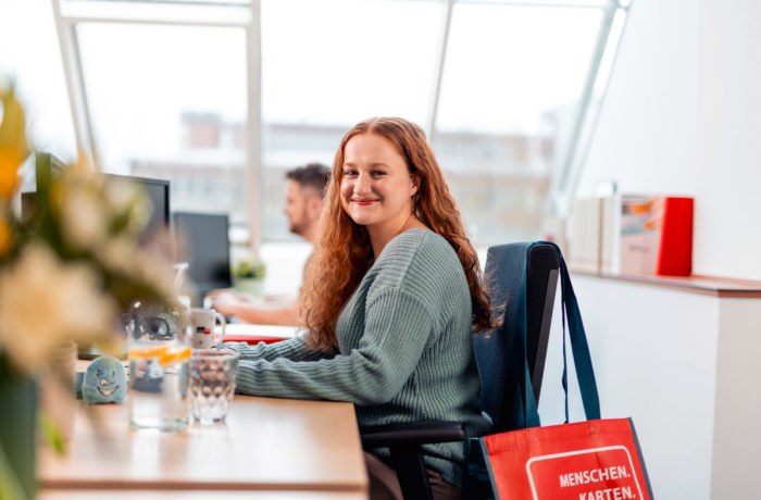 Eine Frau mit langen lockigen Haaren und einem grünen Pullover sitzt an einem Schreibtisch in einem hellen Büro und lächelt in die Kamera. Eine rote Tragetasche hängt an ihrem Stuhl, und im Hintergrund arbeitet eine weitere Person an einem Computer. Eine Frau mit langen lockigen Haaren und einem grünen Pullover sitzt an einem Schreibtisch in einem hellen Büro und lächelt in die Kamera. Eine rote Tragetasche hängt an ihrem Stuhl, und im Hintergrund arbeitet eine weitere Person an einem Computer.