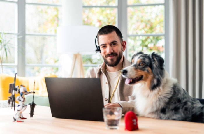 Ein Mann, der ein Headset trägt, lächelt, während er zu Hause an einem Laptop arbeitet. Ein Hund sitzt neben ihm am Tisch. Ein Glas Wasser, ein kleines rotes Spielzeug und ein Robotermodell stehen ebenfalls auf dem Tisch. Im Hintergrund sind helle Fenster zu sehen. Ein Mann, der ein Headset trägt, lächelt, während er zu Hause an einem Laptop arbeitet. Ein Hund sitzt neben ihm am Tisch. Ein Glas Wasser, ein kleines rotes Spielzeug und ein Robotermodell stehen ebenfalls auf dem Tisch. Im Hintergrund sind helle Fenster zu sehen.