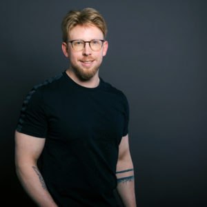 Robert Schwarzmeier, ein Mann mit hellbraunem Haar, einem Bart und einer Brille, steht lächelnd vor einem dunklen Hintergrund. Er trägt ein schwarzes T-Shirt, hat Tattoos auf den Unterarmen und steckt lässig die Hand in die Tasche.