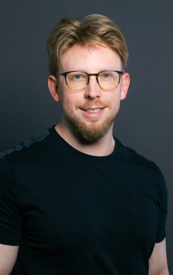 Robert Schwarzmeier, ein Mann mit hellbraunem Haar, einem Bart und einer Brille, steht lächelnd vor einem dunklen Hintergrund. Er trägt ein schwarzes T-Shirt, hat Tattoos auf den Unterarmen und steckt lässig die Hand in die Tasche.