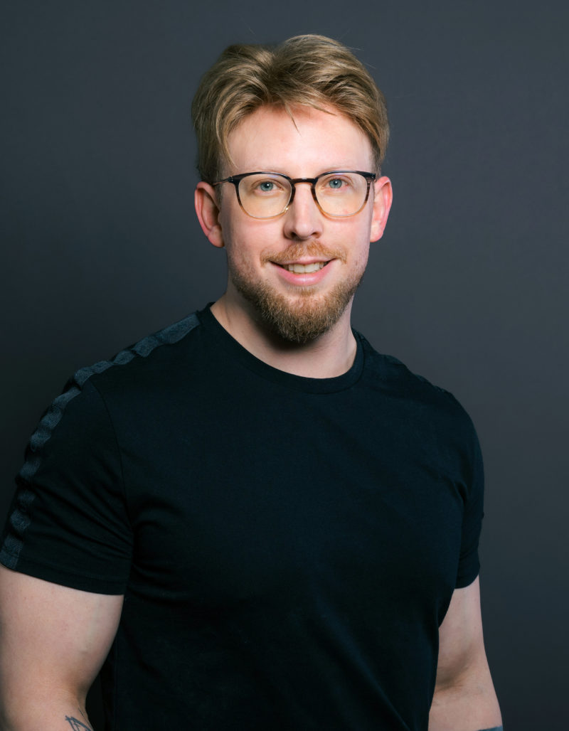 Robert Schwarzmeier, ein Mann mit hellbraunem Haar, einem Bart und einer Brille, steht lächelnd vor einem dunklen Hintergrund. Er trägt ein schwarzes T-Shirt, hat Tattoos auf den Unterarmen und steckt lässig die Hand in die Tasche.