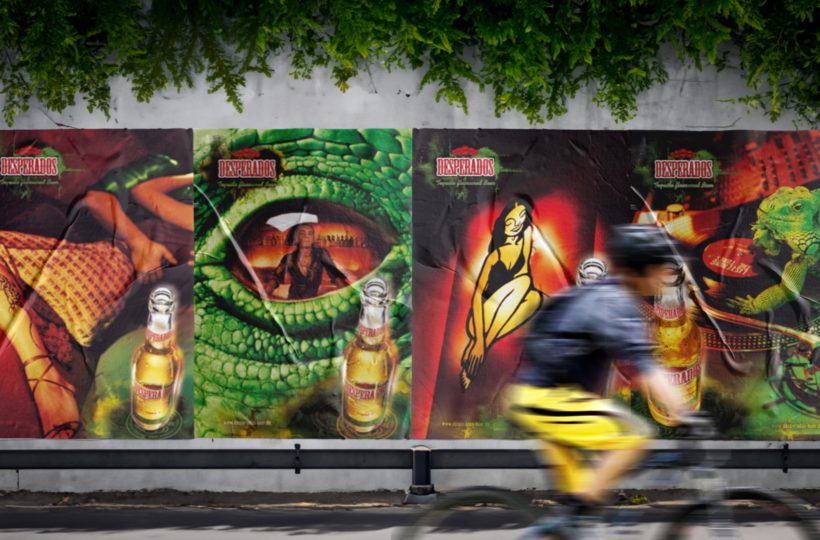 Ein Radfahrer fährt an einer Wand vorbei, die mit farbenfrohen Desperados-Bierplakaten bedeckt ist, die surreale, lebendige Kunstwerke wie ein großes Reptilienauge und stilisierte menschliche Figuren zeigen. Über den Postern hängen Grünpflanzen.