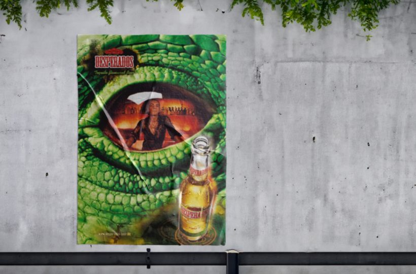 Ein farbenfrohes Plakat mit einer Frau mit Cowboyhut, einer Flasche Desperados-Bier und einem großen grünen Reptilienauge, das auf einer weißen, verwitterten Wand mit darüber hängenden Grünpflanzen zu sehen ist.