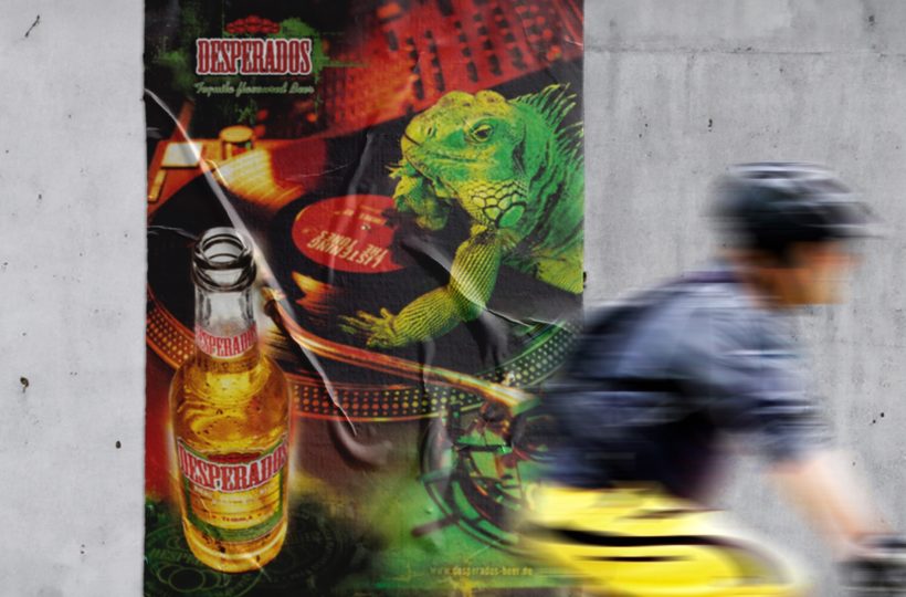 Ein verschwommener Radfahrer fährt vor einem Desperados-Bierplakat vorbei, das einen grünen Leguan mit einem DJ-Plattenspieler, einer Bierflasche und leuchtenden Neonfarben zeigt.