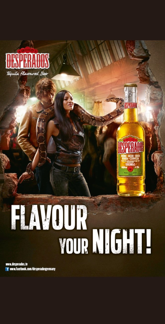 Ein Mann und eine Frau posieren mit einer großen Schlange in einer belebten Bar. Im Vordergrund steht eine Flasche Desperados-Bier. Der Text lautet: Desperados. Flavour your night!.