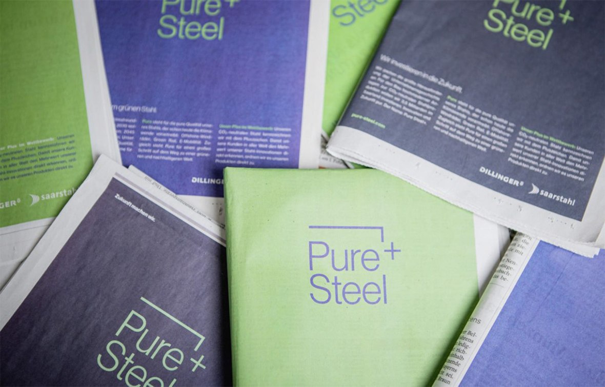 Mehrere Zeitungen mit grünen und blauen Einbänden und dem Titel Pure + Steel in weißer Schrift sind ausgebreitet - ein starker Hinweis auf eine durchdachte Markenentwicklung. Die Seiten enthalten Textblöcke, aber keine Bilder von Menschen.
