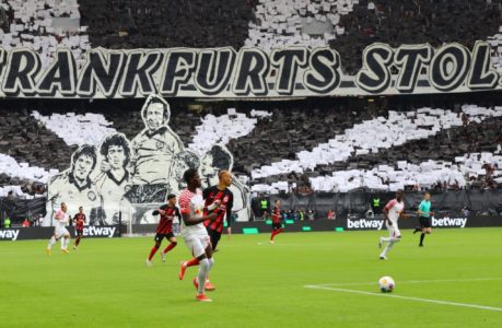 Fußballspieler treten auf einem Feld gegeneinander an, während die Fans auf der Tribüne ein großes Schwarz-Weiß-Display mit Porträts und dem Schriftzug "FRANKFURTS STOLZ" in die Höhe halten.