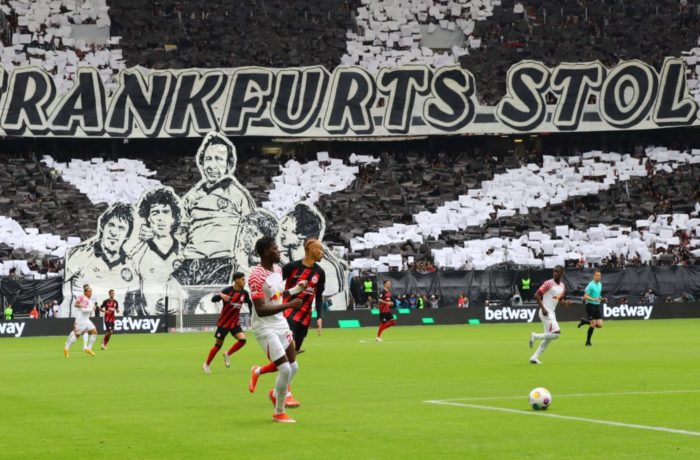 Fußballspieler treten auf einem Feld gegeneinander an, während die Fans auf der Tribüne ein großes Schwarz-Weiß-Display mit Porträts und dem Schriftzug "FRANKFURTS STOLZ" in die Höhe halten.