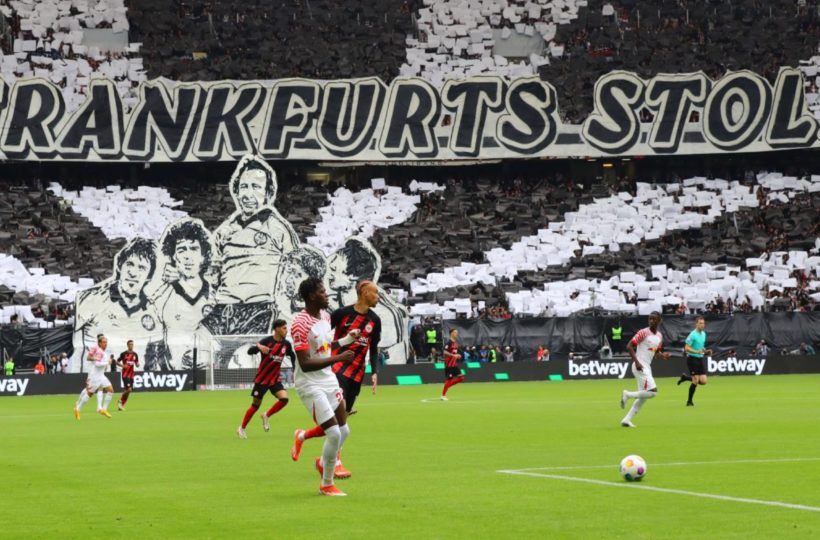 Fußballspieler treten auf einem Feld gegeneinander an, während die Fans auf der Tribüne ein großes Schwarz-Weiß-Display mit Porträts und dem Schriftzug "FRANKFURTS STOLZ" in die Höhe halten.