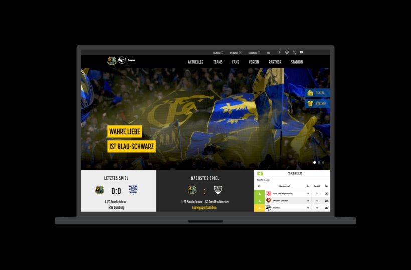 Ein Laptop-Bildschirm zeigt die Website des 1. FC Saarbrücken mit blau-gelben Fahnen im Hintergrund, Spielergebnissen, bevorstehenden Spielen und dem Tabellenstand an. Der Slogan "WAHRE LIEBE IST BLAU-SCHWARZ" ist gelb hervorgehoben.