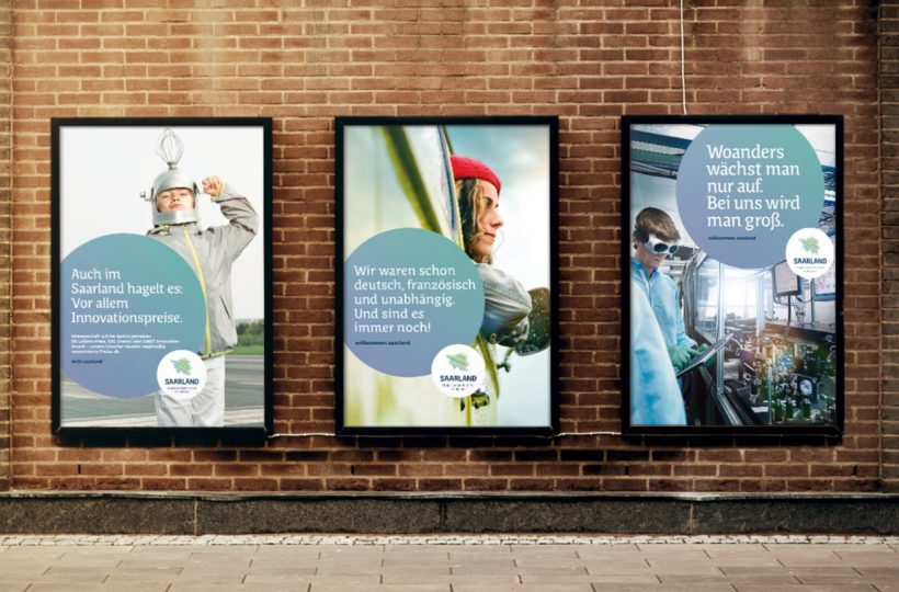 Drei Plakate auf einer Backsteinmauer zeigen Kinder als Erfinder und Wissenschaftler, jedes mit einem Logo des Saarland-Tourismus und einem deutschen Text, der für Innovation, Vielfalt und Wachstum im Saarland wirbt.