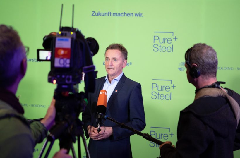 Ein Mann im Anzug spricht während eines Interviews in Mikrofone, während Kameras und Mitarbeiter vor ihm stehen. Der grüne Hintergrund zeigt die Worte Zukunft machen wir. und Pure+Steel.