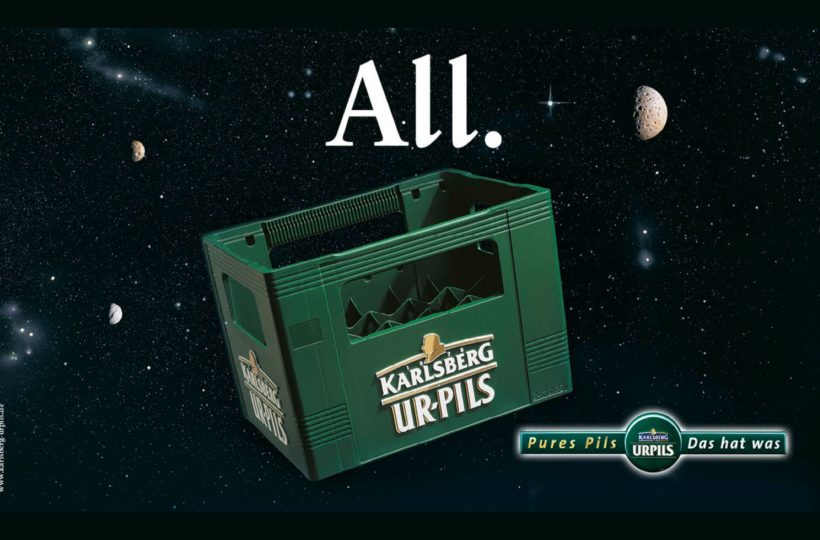 Eine grüne Karlsberg Urpils-Bierkiste schwebt im Weltall, umgeben von Sternen und kleinen Monden, mit dem Wort All. darüber. Das Karlsberg Urpils-Logo und der Slogan erscheinen unten rechts.