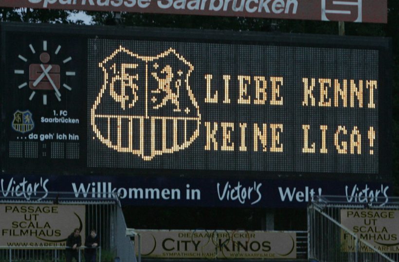 Eine Stadionanzeigetafel zeigt das Emblem des 1. FC Saarbrücken und den deutschen Satz "LIEBE KENNT KEINE LIGA!" mit Werbung darunter und einer Uhr auf der linken Seite.