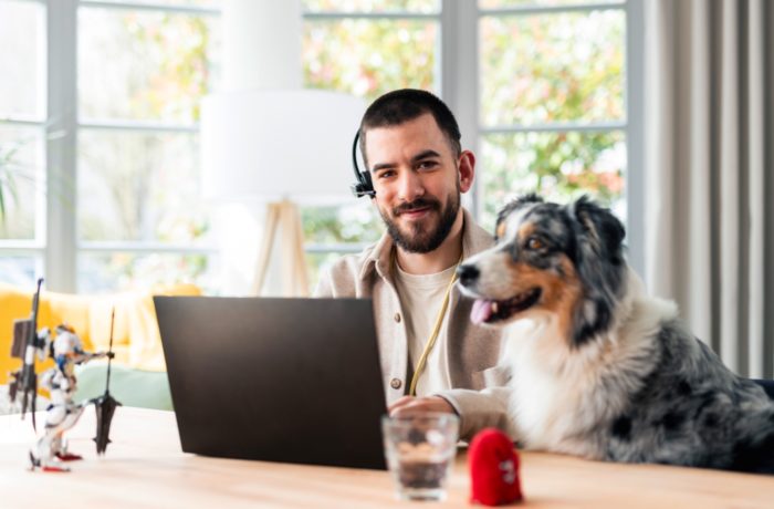 Ein Mann, der ein Headset trägt, sitzt an einem Schreibtisch mit einem Laptop und lächelt in die Kamera. Ein Hund sitzt neben ihm. Auf dem Tisch befinden sich ein Spielzeug, ein Glas Wasser und ein roter Gegenstand. Im Hintergrund sind helle Fenster und Grünflächen zu sehen. Ein Mann, der ein Headset trägt, sitzt an einem Schreibtisch mit einem Laptop und lächelt in die Kamera. Ein Hund sitzt neben ihm. Auf dem Tisch befinden sich ein Spielzeug, ein Glas Wasser und ein roter Gegenstand. Im Hintergrund sind helle Fenster und Grünflächen zu sehen.