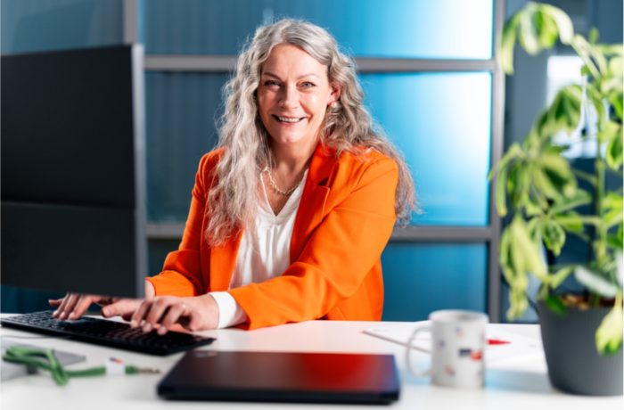 Eine lächelnde Frau mit langen grauen Haaren, die einen orangefarbenen Blazer trägt, sitzt an einem Schreibtisch und tippt auf einer Tastatur. Ein Computermonitor, eine Pflanze und ein Kaffeebecher stehen auf dem Schreibtisch in einer hellen Büroumgebung. Eine lächelnde Frau mit langen grauen Haaren, die einen orangefarbenen Blazer trägt, sitzt an einem Schreibtisch und tippt auf einer Tastatur. Ein Computermonitor, eine Pflanze und ein Kaffeebecher stehen auf dem Schreibtisch in einer hellen Büroumgebung.
