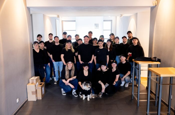 Eine Gruppe von etwa 25 Personen, die meisten mit schwarzen Hemden und Hüten, posiert in einem hellen Innenraum. Marc Hellenthal ist unter ihnen, und zwei Personen knien vorne neben einem Hund. An den Seiten sind Kisten und Tische zu sehen.
