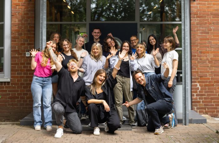Eine Gruppe von sechzehn Personen posiert und lächelt vor einem Backsteingebäude, einige knien davor, andere stehen dahinter. Einige winken enthusiastisch und spiegeln die fröhliche Atmosphäre ihrer Veranstaltung zur Medienkompetenz wider.