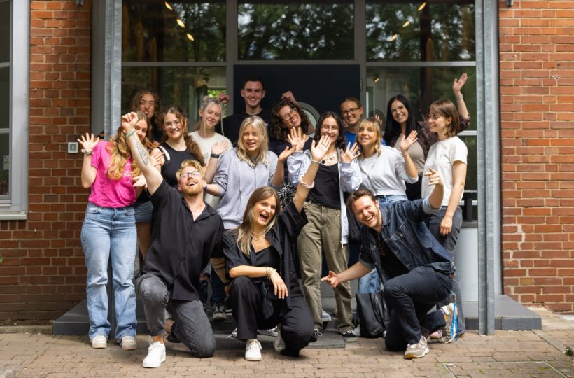Eine Gruppe von sechzehn Personen posiert und lächelt vor einem Backsteingebäude, einige knien davor, andere stehen dahinter. Einige winken enthusiastisch und spiegeln die fröhliche Atmosphäre ihrer Veranstaltung zur Medienkompetenz wider.