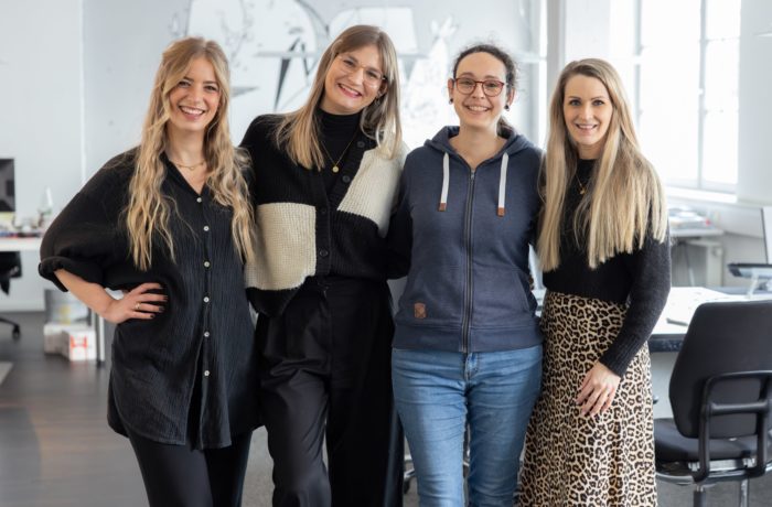 Vier neue Ausbilderinnen stehen dicht beieinander und lächeln in die Kamera in einem hellen, modernen Büroraum. Drei sind schwarz gekleidet, eine trägt einen blauen Kapuzenpulli und Jeans. Im Hintergrund sind Schreibtische und Bürostühle zu sehen.