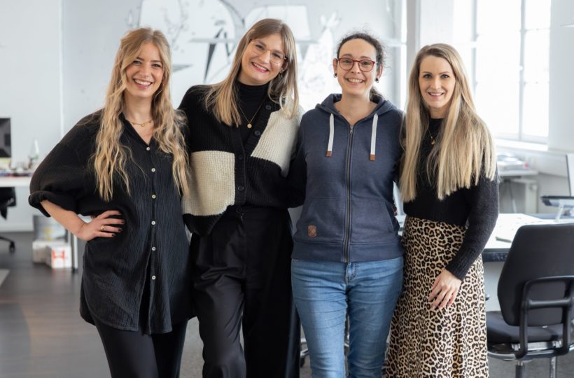 Vier neue Ausbilderinnen stehen dicht beieinander und lächeln in die Kamera in einem hellen, modernen Büroraum. Drei sind schwarz gekleidet, eine trägt einen blauen Kapuzenpulli und Jeans. Im Hintergrund sind Schreibtische und Bürostühle zu sehen.