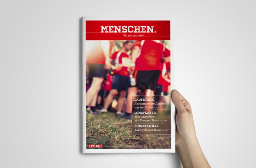 Eine Hand hält eine Zeitschrift mit einem Titelbild, das Menschen in roten Hemden beim Laufen zeigt, mit dem Titel MENSCHEN. in fetter weißer Schrift auf rotem Hintergrund und mehreren darunter aufgeführten Artikelüberschriften.