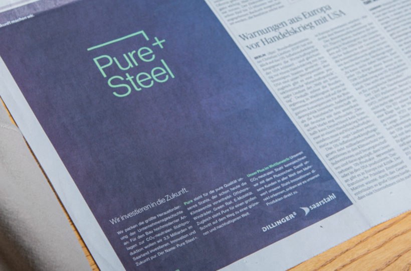 Eine Zeitungsseite zeigt eine große Anzeige mit dem Text Pure + Steel in Grün auf dunklem Hintergrund. Darunter befinden sich ein deutscher Text und Logos für Dillinger und Saarstahl. In den angrenzenden Spalten stehen die üblichen Nachrichtenartikel in deutscher Sprache.