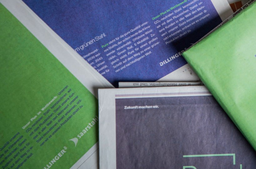 Nahaufnahme von mehreren sich überlappenden Zeitungen mit grünem und blauem Einband und deutschem Text, der teilweise sichtbar ist. Der Text enthält Schlagzeilen und Firmennamen.